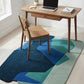 Wooden desk with laptop and chair on a colorful rug in a bright room.