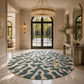 A large round area rug with an abstract organic pattern in teal blue and cream, styled in a luxurious, high-end entryway with marble floors, a gold chandelier, and a console table.