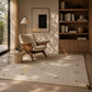 A large cream-colored area rug with subtle geometric patterns is styled in a warm, modern living room with a mid-century armchair, wooden bookshelf, and natural sunlight filtering through a window.