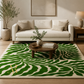 A large area rug with a bold green and cream abstract leaf-like pattern, styled in a modern living room setting with a cream sofa, wooden coffee table, and potted plant.