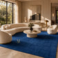 A rectangular royal blue textured rug with a small gold embroidered monogram logo in the upper right quadrant, shown flat against a plain white background.