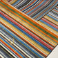 A close-up of a striped area rug with multiple colors including blue, orange, green, red, yellow, beige, and gray in diagonal bands, with a gold embroidered letter 'R' on the lower left.