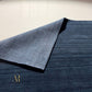 A close-up of a dark navy blue textured rug with a folded corner showing the underside, placed on a light beige tiled floor. A small gold 'AR' logo is visible in the lower-left corner of the rug.