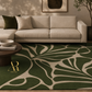 A green and beige abstract leaf-patterned area rug styled in a modern living room setting with a beige sectional sofa, a dark wood coffee table, and neutral-toned walls.