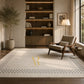 A large cream-colored area rug with a black geometric border and a gold monogram logo in the center, styled in a modern, warm-toned living room next to a mid-century armchair and a built-in wooden bookshelf.
