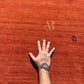 A close-up of a textured orange-red rug with a person's hand placed on it for scale, along with two embroidered
and a gold 'AR'