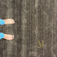 A close-up of a dark brown and gray textured area rug with vertical heathered stripes, shown from above with a person's bare feet and blue jeans visible on the left side. A gold 'R' logo is printed in the lower right corner.