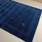 A dark navy blue rectangular rug with a textured surface, featuring a decorative border of small white dots and a gold monogram logo in the center, shown against a plain white background.