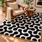 A black and cream geometric patterned area rug styled in a bright, modern living room next to a wooden armchair and a small side table.