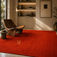 A bright red area rug with a gold monogram logo in a modern, sunlit living room featuring a wooden lounge chair, shelving, and a floor lamp.
