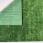Two textured green fabric or material swatches layered at a corner, with a small gold monogram logo near the intersection, shown on a white background.