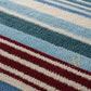 Close-up of a textured knit fabric with horizontal stripes in light blue, teal, cream, and burgundy, featuring a small gold logo watermark in the lower right.