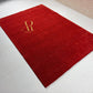 A red rectangular area rug with a gold monogram logo reading 'AR' and 'Arts Rug' in the upper left, photographed on a light tiled floor.