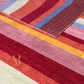 A close-up of a striped area rug with textured horizontal bands in cream, red, pink, yellow, orange, and blue, featuring a gold monogram logo.