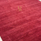 A close-up of a deep burgundy red textured rug with a subtle heathered pattern and a small gold embroidered monogram logo near the center.