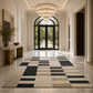 A large rectangular area rug with a modern geometric patchwork design in beige, cream, and black, displayed in a luxurious, bright entryway with marble floors and a chandelier.