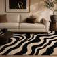 A modern living room scene featuring a black and beige wavy patterned area rug on the floor, a cream-colored fabric sofa with brown accent pillows, a dark wood coffee table, and a framed art print on the wall.