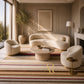 A styled interior scene with a cream boucle sofa, two matching armchairs, a round stone coffee table, a striped area rug, and a tall potted tree in a warm, sunlit room.
