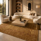 A large textured area rug in warm brown and gold tones with a stylized gold monogram, styled in a modern living room with cream curved sofas, a wooden coffee table, and warm ambient lighting.