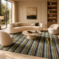 A large rectangular area rug with vertical stripes in navy, green, beige, and muted gold, styled in a luxurious modern living room with a curved cream sofa, two matching armchairs, and a round stone coffee table.