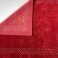 A red rectangular rug with a decorative border of small white dots and a gold monogram in the upper right corner, shown flat against a white background.