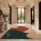 A large abstract area rug with organic shapes in teal, terracotta, blush pink, and cream, displayed on a polished marble entryway floor. The rug features a gold monogram logo in the bottom-left corner.