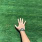 A hand and forearm resting on a bright green artificial turf or grass-like surface, with a wristwatch visible. The hand is placed to show scale on the textured green surface.