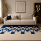 A modern living room scene featuring a cream-colored sofa with two accent pillows, set on a large cream rug with a bold blue circular pattern. The room has beige walls, a wooden bookshelf, and a potted plant.