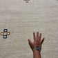 A rectangular beige area rug with a Southwestern geometric pattern in brown, gold, and black, featuring a gold monogram logo near the bottom right. The rug is shown flat against a solid white background.