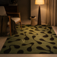 A rectangular area rug with an abstract organic wavy pattern in olive green and black, styled in a warm, modern living room setting with a wooden armchair, a floor lamp, and a small side table.