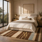A styled bedroom interior with a large striped area rug in brown, beige, cream, and muted blue tones. The rug features a prominent gold monogram logo reading 'RR' with 'Royal Rugs' beneath it.