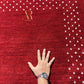 A close-up of a deep red textured rug with a gold 'Armani-Rugh' logo in the upper left and small white floral motifs along the top and right edges. A person's hand wearing a watch rests on the rug to show scale.