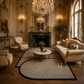 An ornate classical living room with cream upholstery furniture, gold-trimmed chandeliers, a marble coffee table, and a large black-and-cream striped area rug on a herringbone wood floor.