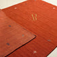 A close-up of a rust-orange textured rug with small scattered metallic emblem motifs and a gold 'AR' monogram near the center, shown on a light neutral floor with a folded section revealing the pile direction.