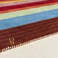 A close-up of a striped wool rug with a thick brown border, cream twisted fringe, and a gold embroidered monogram logo on the corner, shown on a light floor.