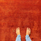 A top-down view of bare feet wearing light blue jeans standing on a textured rust-red rug with a small gold monogram near the left foot.