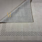 A light beige area rug with a geometric black dotted border pattern, shown with one corner folded over to display the texture and underside.