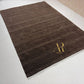 A dark brown rectangular area rug with a subtle heathered texture, featuring a gold embroidered monogram logo reading 'ARANDA RUGS' in the bottom right corner, displayed on a light-colored tiled floor.