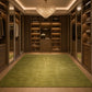 A luxury walk-in closet with dark wood cabinetry, mirrored doors, and a large green area rug featuring a gold monogram logo. A crystal chandelier hangs from the ceiling, and shoes are neatly displayed on shelves.