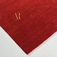 A close-up of a deep red textured rug or fabric sample with a gold 'AR' monogram logo in the upper left, placed on a light gray surface.