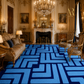 A large blue and navy geometric patterned area rug displayed in an ornate, classical European-style living room with gold-trimmed walls, a fireplace, chandeliers, and vintage furniture.