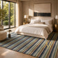 A styled bedroom interior featuring a large striped area rug in muted blue, green, beige, and brown tones. The rug is laid out on a light wood floor in front of a bed with white linens, flanked by gold bedside tables and lamps, with a large window showing greenery outside.