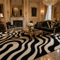 A luxury living room featuring a large black and beige wavy patterned area rug. The room is styled with elegant gold-framed furniture, including a black velvet sofa with gold trim, a matching armchair, and a marble coffee table.