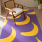 A purple area rug with a bold yellow banana pattern, styled in a bright room with a wooden armchair and a potted plant.