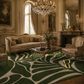 An ornate classical living room featuring a large green and beige abstract leaf-patterned area rug. The room is decorated with gold-trimmed walls, a crystal chandelier, a dark wood fireplace, and matching vintage upholstered furniture.