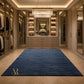 A large rectangular navy blue area rug with a gold monogram logo in the bottom-left corner, displayed in a luxurious, high-end walk-in closet with wood floors and built-in cabinets.
