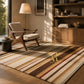 A striped area rug in warm beige, brown, cream, and muted red tones, styled in a modern living room with a mid-century armchair, wooden bookcase, and floor lamp.
