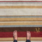 A top-down view of a striped area rug with beige, brown, red, and blue-gray horizontal bands, with bare feet standing on the rug and a gold 'AR Anita Ring' logo in the lower right corner.