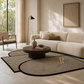 A modern living room scene featuring a large, stylish area rug with organic wavy shapes, black and beige stripes, and a gold monogram. The rug is styled in a warm, minimalist room with a cream-colored sofa, a wooden coffee table, and a black floor lamp.