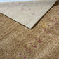 A close-up of a woven area rug showing its texture and pattern. The rug features a warm tan and brown base with a subtle heathered effect, accented by small repeating dusty rose or mauve geometric diamond motifs.